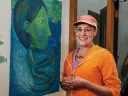 A woman in an orange sweater and cap stands in front of a large blue and green painting of a water-skiing woman.