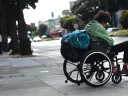 A man in a wheelchair with a rolled-up sleeping bag strapped to the back