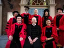 8 people in academic robes sit for a photograph in front on Memorial Church