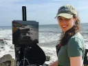 Hudson in a baseball cap with her easel and paints at a rocky seaside