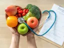 Hands holding a heart-shaped bowl of fruits and vegetables with a stethoscope, clipboard with medical records in the background.