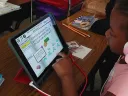 A young girl with pink headphones uses a tablet for an educational reading game at a classroom desk.