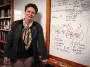 Catherine Dulac stands beside a whiteboard filled with colorful handwritten notes and diagrams, wearing a dark blazer and patterned scarf.