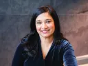 Moitri Chowdhury Savard smiling, sitting indoors against a dark background.