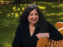Jane Rosenzweig seated on a wooden bench in a grassy outdoor setting, smiling warmly.