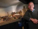 Frank Gevalt standing beside his detailed model city "Port Charles," featuring buildings, bridges, and an urban skyline.