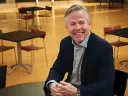 Tom Kane smiles  in a navy blazer and sits at a table in a modern room with empty chairs and tables behind him.