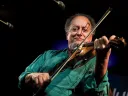 A violinist in a green shirt plays on stage, smiling, with microphones and stage lighting in the background.