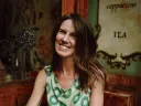A person with long brown hair wears a sleeveless green dress with a kiwi fruit pattern, sitting in a vintage-style café.