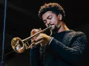 Toussaint Miller  in a dark sweater plays the trumpet on stage with eyes closed.