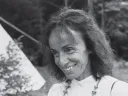 A black-and-white portrait of a smiling person with wavy hair, wearing a beaded necklace.