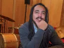 Jesse Cox smiling at a table with music notes and a book, with drums and cymbals behind him
