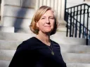 Hopi Hoekstra standing on outdoor steps in sunlight, wearing a dark top and pendant necklace