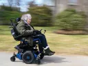 Lisa Iezzoni smiling while riding a motorized wheelchair on a paved path through a grassy area