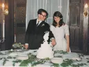 A bride and groom in formal attire cut a tiered cake at a candlelit wedding reception table.