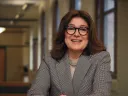 Nicolo Maestas in a grey suit and wearing glasses sits with her arms on a table
