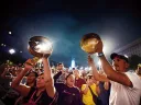 A crowd of people shout and march during a nighttime demonstration, while a man and woman in the foreground hold two silver-colored pans above their heads and bang on them with sticks