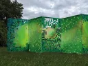 A six-foot-tall, five-panel folded screen stands in a field of grass next to the woods. It's painted different shades of green, with some squares cut out that represent digital pixels.