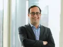 Matt Levine in a dark blazer and glasses stands smiling with arms crossed in front of a large window in a city building.