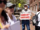 Man, standing in small group of people outside the courthouse, holding a sign that reads "HANDS OFF HARVARD" in red letters