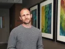 Man in gray sweater standing in hallway with colorful abstract art on wall.