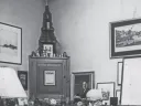 David McCord in suit reading a book at cluttered wooden desk in office filled with framed art and shelves.