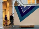 Two people moving large abstract painting with blue V-shaped design in museum courtyard.