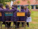 Students in purple jackets seated on chairs, facing away in a grassy area.