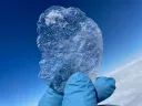 A gloved hand holds a clear shard of blue ice against the sky.