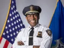 Smiling police chief in uniform, standing with arms crossed between American and state flags.