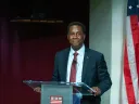 Setti Warren stands at a podium, smiling, with an American flag in the background.