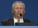 Lawrence Summers in a suit  at a podium with a microphone, looking serious.