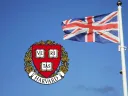 Union Jack flag waving next to the Harvard University crest against a blue sky.