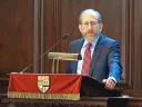 A man in a suit speaking at a podium with a red drape and a university crest.