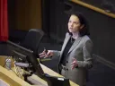A woman in a gray suit gestures while speaking at a podium in a lecture hall.