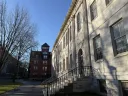 University Hall building with steps, shadows of trees, and a red brick structure in the background.