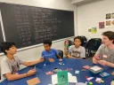 Four young people sitting around a table playing a card game, with a chalkboard in the background.