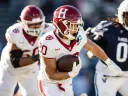Harvard football player runs with the ball, pursued by opposing players on the field.