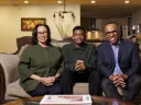 Nancy Hill, Rendall Howell, and their son Theo sit together on a couch in a cozy living room, smiling at the camera.