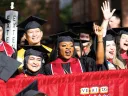 Graduates celebrate joyfully, wearing caps and gowns, with some waving and smiling.