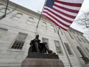 A seated statue of a man reading, with an American flag waving in the background.