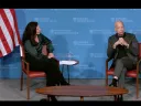 A discussion panel featuring two speakers seated in chairs, with a blue backdrop and an American flag.