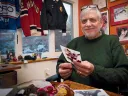 Older man in a green sweater holds a postcard in a warmly decorated office.
