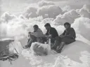 Three climbers seated on a snowy summit, surrounded by clouds, appearing contemplative.