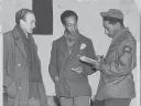 Three men engaged in conversation, one taking notes