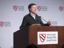 A speaker gestures while presenting at a podium with Harvard Radcliffe Institute branding.