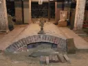 Brick archway with a sandy base, surrounded by wooden planks and boxes in a dim space.