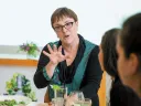 A woman in glasses gestures while speaking to two attentive listeners at a table.