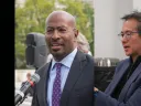Two men in professional attire, one speaking at a microphone, the other gesturing while talking.