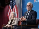 Two figures sitting against an American flag having a conversation at Harvard Kennedy School